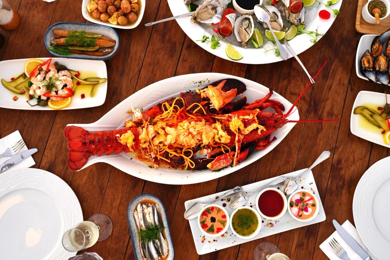 Masa üstünde, ortada kırmızı kabuklu, kısmen açılmış ve spagetti ile servis edilmiş büyük bir ıstakoz tabağı yer alırken, çevresinde istiridye, midye, karides salatası, hamsi, patates ve çeşitli mezelerle donatılmış zengin bir deniz ürünleri ziyafeti kuşbakışı sergileniyor.