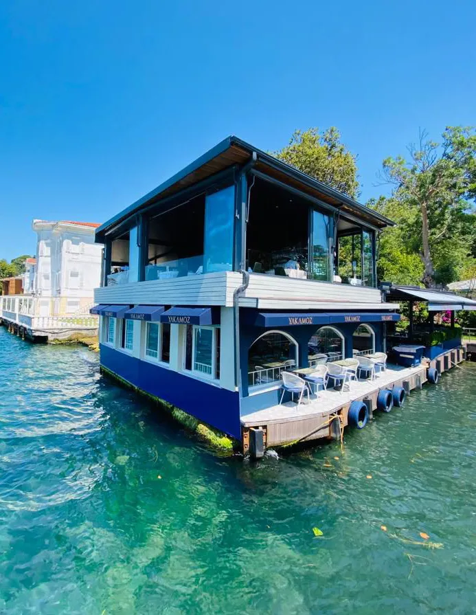 Berrak turkuaz sular üzerinde yer alan, alt katı lacivert, üst katı büyük cam pencerelerle çevrili, YAKAMOZ yazılı tenteleri olan iki katlı modern bir restoranın güneşli bir günde çekilmiş genel görünümü; restoranın önündeki ahşap iskelede boş masa ve sandalyeler seçilirken, arka planda beyaz bir yalı ve yemyeşil ağaçlar görülüyor.