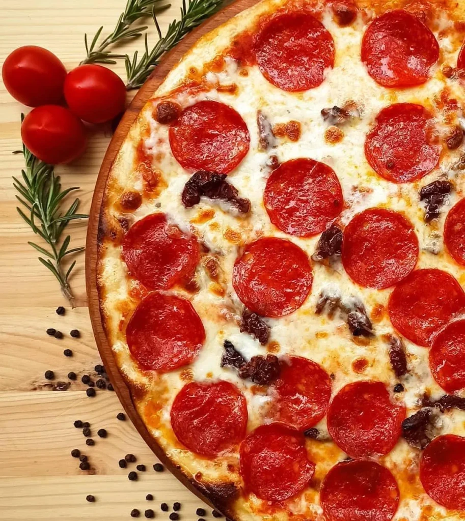Ahşap bir zeminde, parlak kırmızı sucuk dilimleri ve erimiş beyaz peynirle kaplı, kenarı hafif kızarmış, iştah açıcı bir 400 derece pizza; yanında üç adet çeri domatesi, biberiye dalları ve taneli karabiberler duruyor.