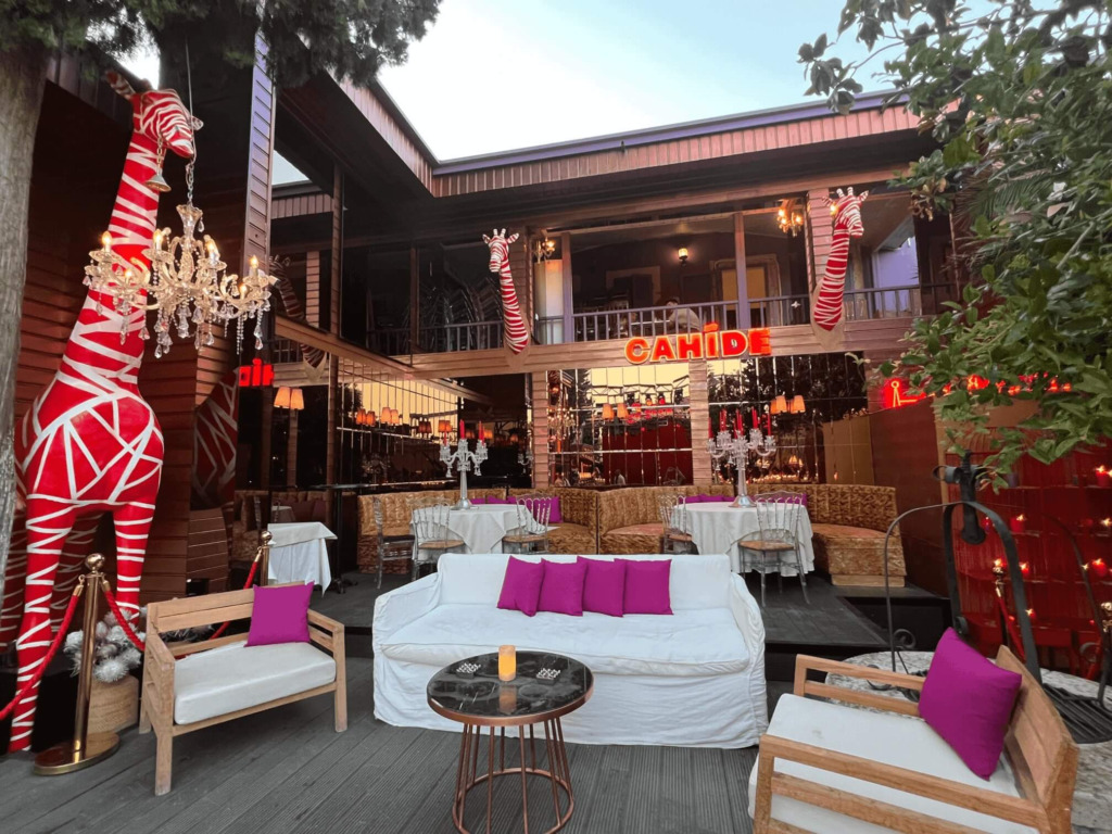 Çeşme’nin hareketli gece mekanlarından Cahide Alaçatı’nın ahşap cepheli, kırmızı beyaz çizgili zürafa heykelleri, kristal avizeler ve fuşya yastıklarla dekore edilmiş şık dış oturma alanında, beyaz kanepeler ve masalar geceye hazır, davetkar bir ambiyans sunuyor.