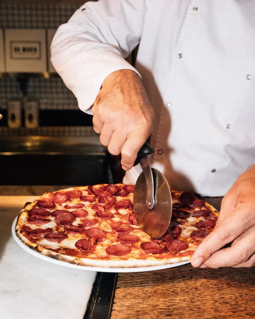 Aşçı şef üniforması giymiş bir adam, arka planda bir restoran mutfağının seçildiği bir ortamda, üzerinde bol sucuk ve erimiş peynir bulunan taze pişmiş yuvarlak bir pizzayı parlak metal bir kesiciyle dikkatlice dilimlerken görülüyor.