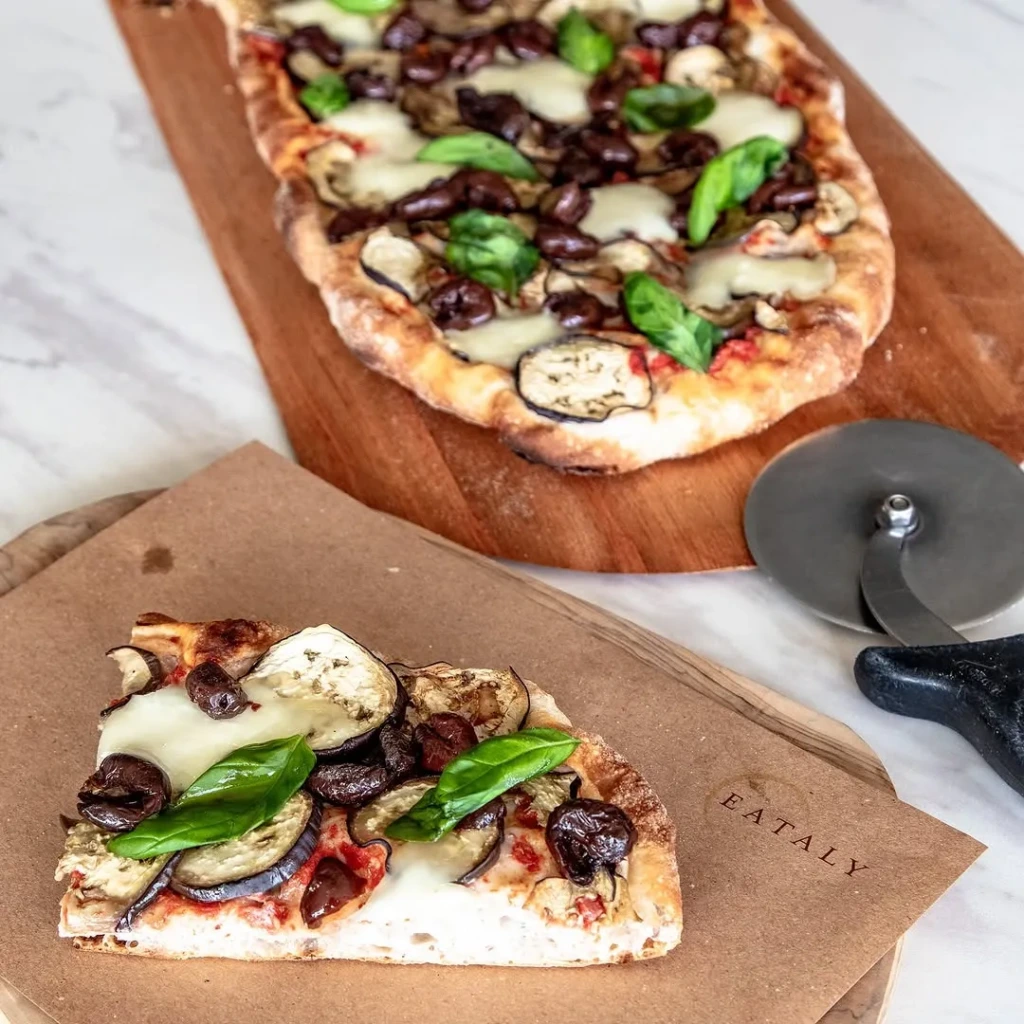Eataly markalı kahverengi kağıt üzerinde duran, patlıcan dilimleri, siyah zeytinler, erimiş mozzarella peyniri ve taze fesleğen yapraklarıyla zenginleştirilmiş bir pizza dilimi, yanında metal saplı parlak bir pizza kesiciyle birlikte ahşap bir servis tahtasında yer alıyor; arka planda ise aynı pizzanın oval tümü mermer zemin üzerinde duruyor.