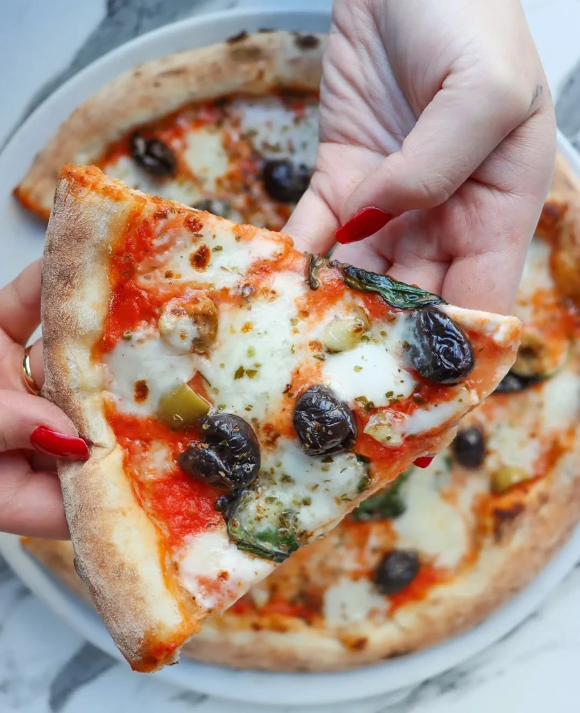 Kırmızı ojeli bir elin özenle tuttuğu, taze pişmiş, kenarları hafif yanık Napoli usulü pizzanın bir dilimi erimiş beyaz mozzarella peyniri, parlak kırmızı domates sosu, siyah ve yeşil zeytinler ile fesleğen yapraklarıyla doluyken, arka planda bulanık bir şekilde başka bir bütün pizza görünüyor.