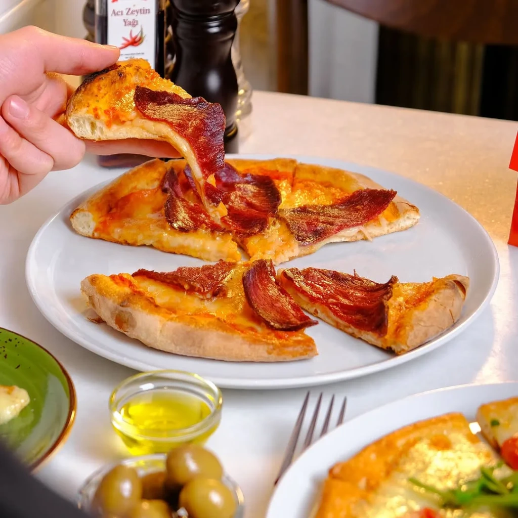Beyaz bir tabakta duran, üzeri bol erimiş peynir ve pastırma benzeri koyu kırmızı et dilimleriyle dolu bir Forno Balat pizzasından bir dilimi kaldıran el, uzayan peynirin iştah açıcı görüntüsünü sergiliyor; arka planda acı zeytinyağı şişesi ve biber değirmeni, ön planda ise zeytinler ve zeytinyağı kasesiyle bir ziyafet masası görünüyor.