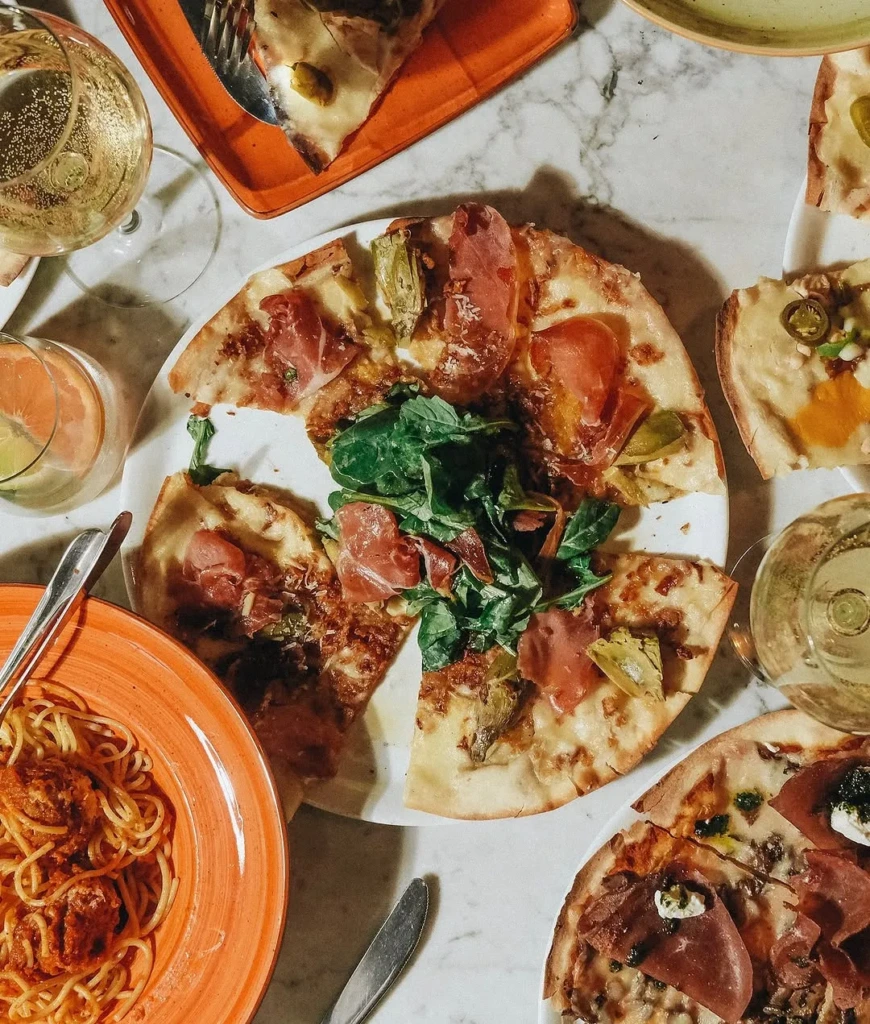 Kuşbakışı bir çekimde, İmperia Ristorante'de mermer bir masanın üzerinde, ortasında roka, enginar ve proşutto dilimleriyle süslenmiş, yarısı yenmiş bir pizza, yanında köfteli spagetti tabağı, iki kadeh köpüklü şarap ve narenciyeli su gibi çeşitli İtalyan yemekleri ve içecekleri ile dolu canlı bir ziyafet sofrası görülüyor.