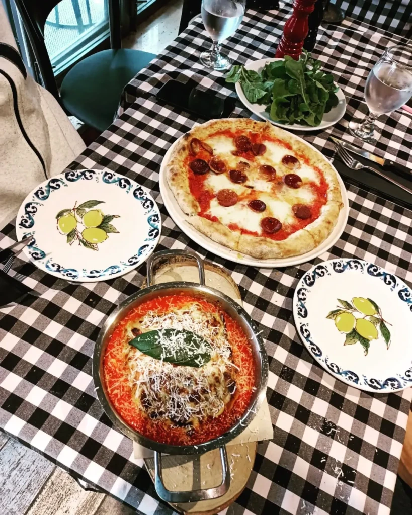 Siyah beyaz kareli masa örtüsü serili bir masada, bol sucuklu ve erimiş mozzarella peynirli geleneksel Napoliten pizza, fırında pişmiş bol parmesanlı domates soslu bir ana yemek, taze roka salatası ve dekoratif zeytin motifli tabaklarla donatılmış lezzetli bir İtalyan gurme sofrası manzarası.