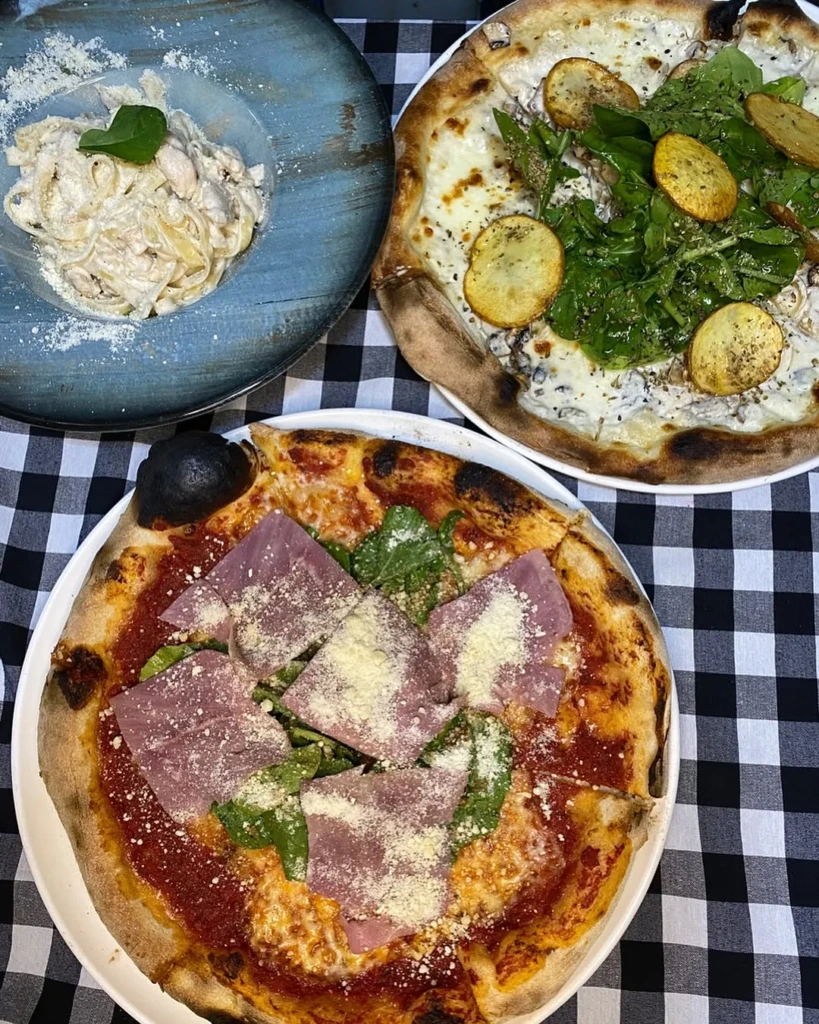 Napoli gourmet restoranında sunulan göz alıcı bir masada, siyah beyaz kareli masa örtüsü üzerinde; kremalı tavuklu fettuccine, beyaz soslu mantar ve patates dilimli taze rokalı odun fırını pizzası ile domates soslu jambon ve peynirle zenginleştirilmiş diğer bir Napoli pizzası iştah açıcı şekilde duruyor.
