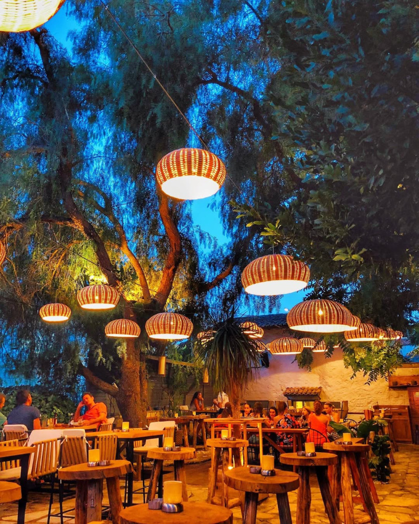 Çeşme'nin en iyi happy hour ve gece mekanları arasında yer alan, büyük bir ağacın altına kurulmuş açık hava ortamında, şık hasır sarkıt lambaların aydınlattığı masalarda sohbet edip içeceklerini yudumlayan insanlar.