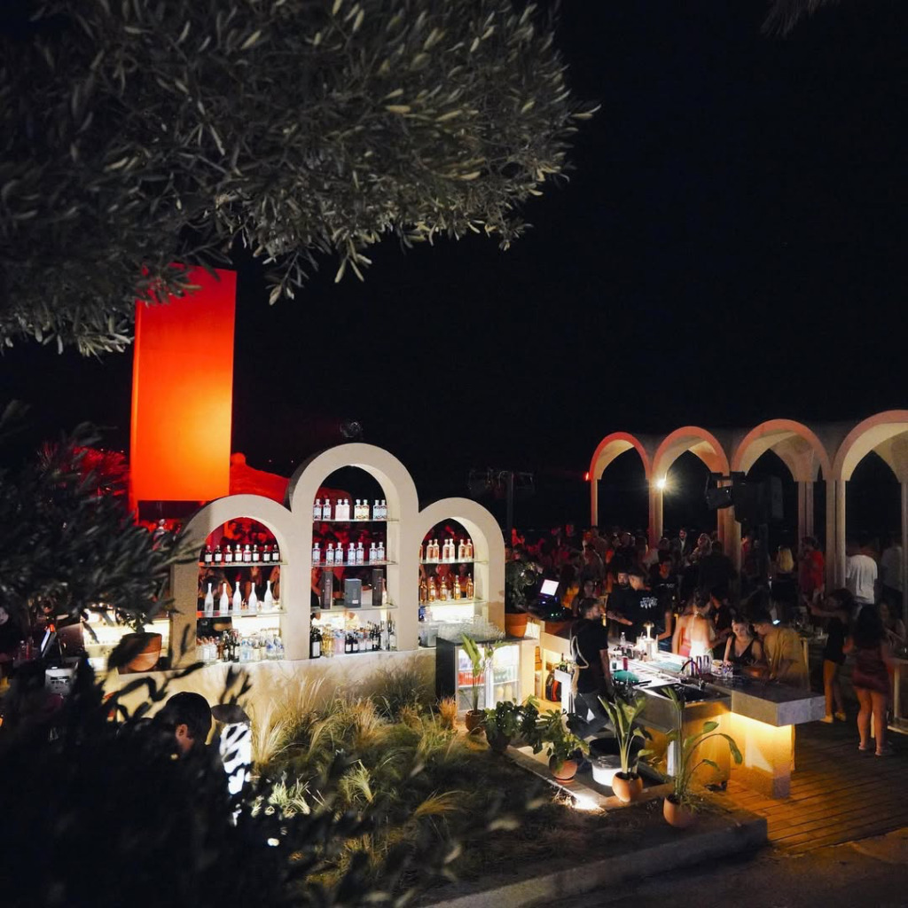 Kemerli beyaz raflarla dolu ada şeklindeki barın önünde DJ eşliğinde dans eden kalabalığın tropik bitkiler ve uzaktaki kemerli yapılar arasında kırmızı ışıklarla aydınlatıldığı, Çeşme'nin enerjik happy hour ve gece mekânlarından Isla'dan hareketli bir akşam görünümü.