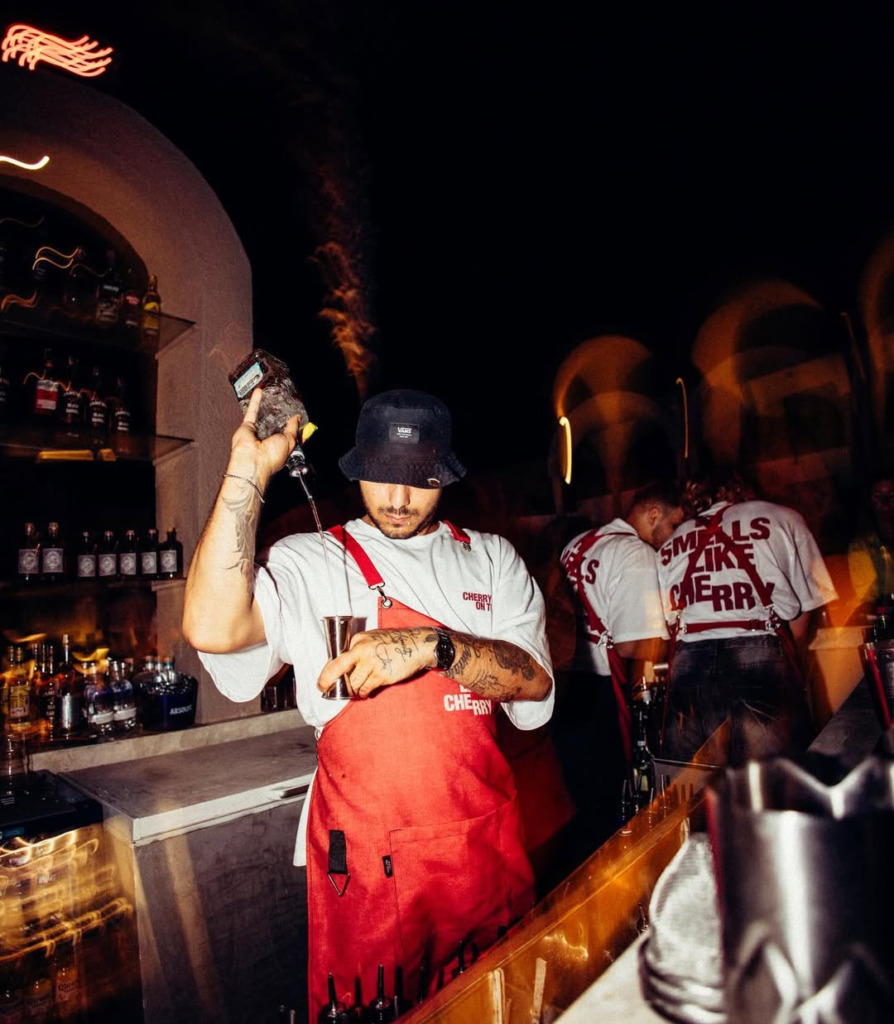 Çeşme’nin hareketli gece mekanlarından Cherry On Top’ta, Vans şapkalı ve dövmeli bir barmen kırmızı önlüğüyle iddialı kokteyller hazırlarken, arka planda bulanık ışıklar ve kalabalık happy hour eğlencesinin enerjisini yansıtıyor.