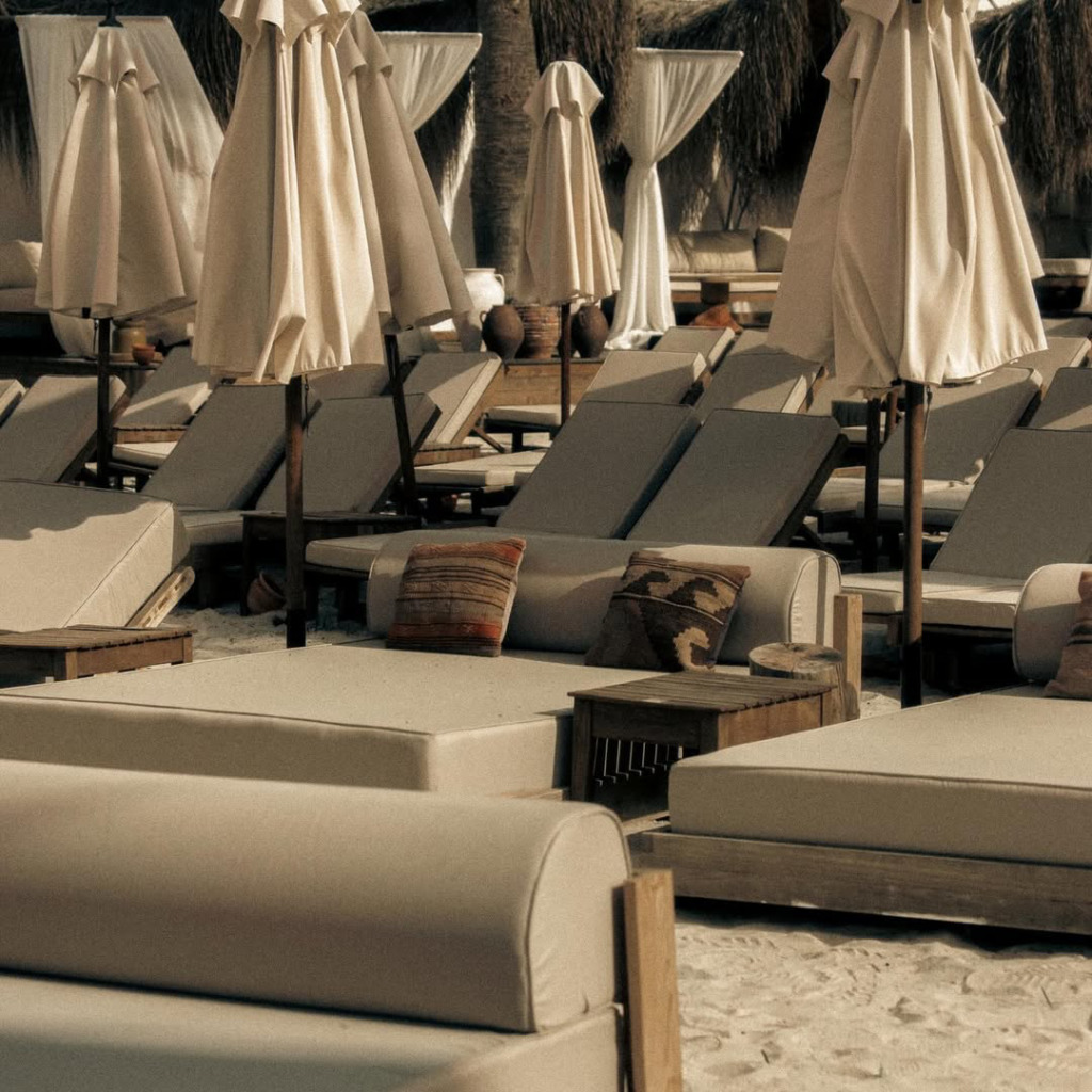 Çeşme ve Alaçatı'daki lüks beach club'lardan birinde, bej renkli konforlu şezlonglar, kapalı plaj şemsiyeleri ve doğal tonlardaki dekoratif yastıklarla bezenmiş, sakin ve huzurlu bir atmosfer sunan kumlu bir dinlenme alanı görülüyor.