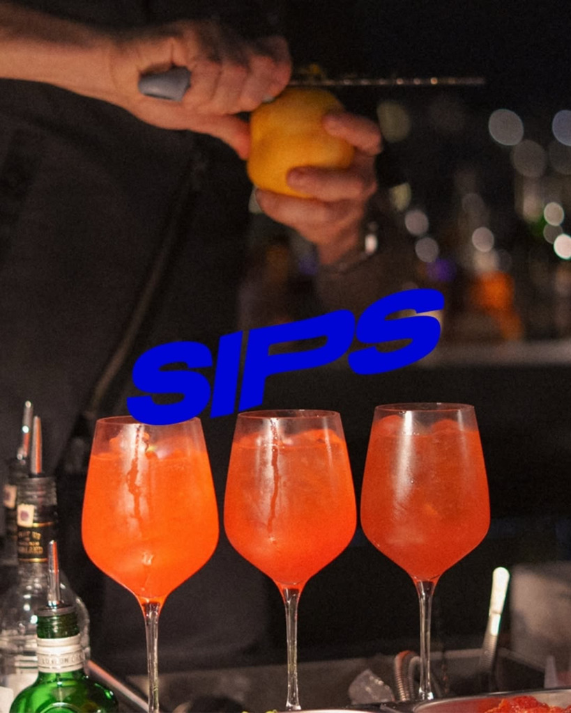 Bir barmen, üç kadeh içindeki turuncu kokteyllerin üzerine bir limonu zester ile soyarak garnitür hazırlarken, Sips Bodrum'un hareketli gecelerindeki özel kokteyl deneyimini yansıtan canlı bir an.
