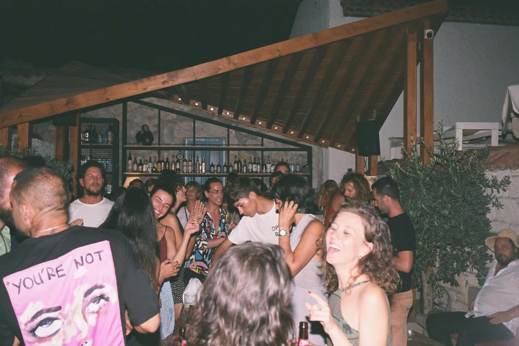 Çeşme’nin en iyi gece mekanlarından biri olan Isla Alaçatı'da, loş ışıklı ve ahşap detaylarla bezeli açık alanda, canlı müzik eşliğinde neşeyle dans edip sohbet eden kalabalık bir grup insan, bar tezgahında sıralanmış çeşitli içeceklerle hareketli bir happy hour deneyimi yaşarken görülüyor.