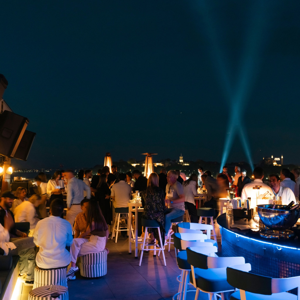 Gece vakti, İstanbul'un en iyi teraslı mekanlarından birinde, mavi ışıklı modern bir bar çevresinde sohbet eden kalabalık misafirler ve uzakta belirginleşen tarihi yarımada silüeti ile gökyüzünü aydınlatan güçlü ışık huzmeleri, şehrin canlı yaz akşamlarını vurguluyor.