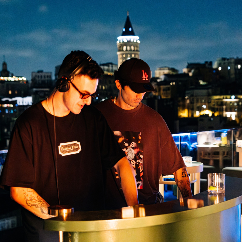 İstanbul'un canlı gece hayatını yansıtan bu teraslı mekanda, kulaklıklı bir DJ ve yanında bir yardımcı, ışıklı bir barın arkasında müzik setini ayarlarken arkalarında Galata Kulesi ve şehrin ışıltılı manzarası belirginleşiyor.