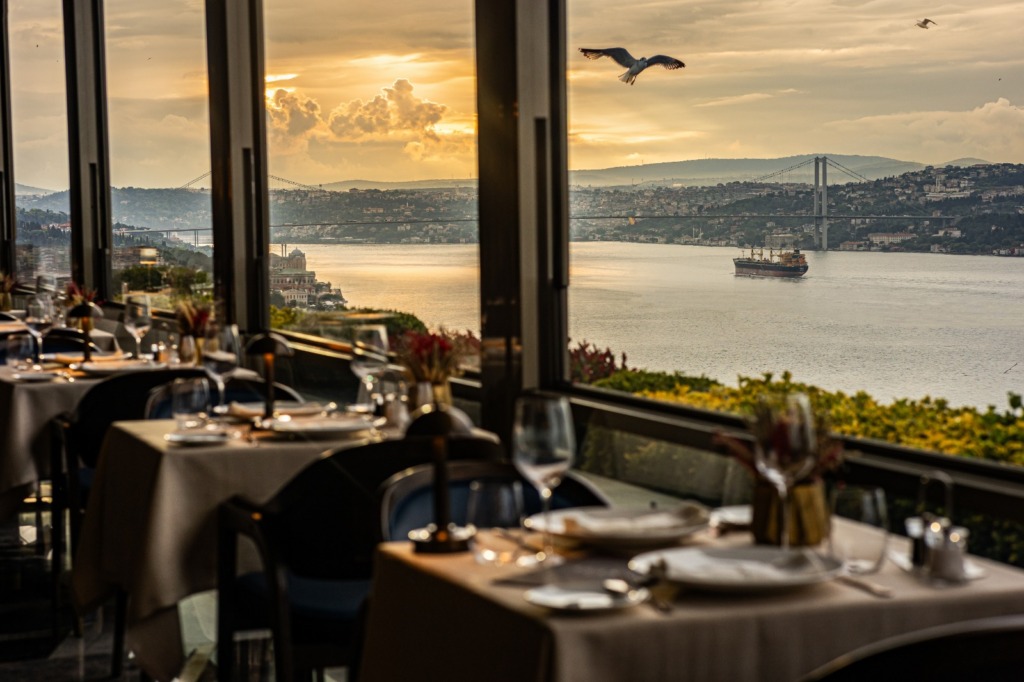 İstanbul’un eşsiz Boğaz ve Köprü manzarasına sahip, zarifçe hazırlanmış masaların ve kadehlerle dolu şık bir restoranın geniş pencerelerinden görünen, gün batımında martıların süzüldüğü ve bir yük gemisinin geçtiği anın fotoğraflandığı bu kare, şehrin en iyi teraslı mekanlarındaki unutulmaz bir deneyimi çağrıştırıyor.