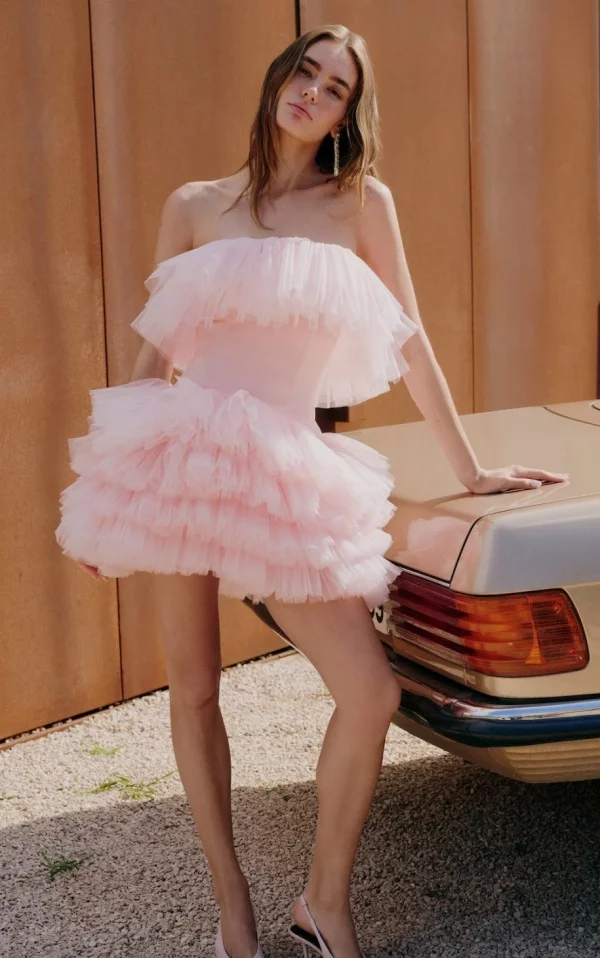Açık pembe, kat kat fırfırlı, straplez mini elbise giyen genç bir kadın, sol elini bej rengi, üstü açılır klasik bir otomobilin arka kısmına zarifçe yaslamış; uzun dalgalı saçları, parlak küpeleri ve açık renk topuklu ayakkabılarıyla çakıllı bir zeminde poz verirken doğrudan kameraya bakıyor.