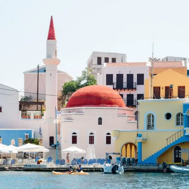 Meis Adası'nın canlı limanında, kırmızı kubbeli cami ve ince minaresiyle öne çıkan rengarenk binaların arasında, suda kayıklarla gezinen insanların keyifli bir anını yansıtan bu kare, adanın görülmeye değer yerlerinden birini gözler önüne seriyor.