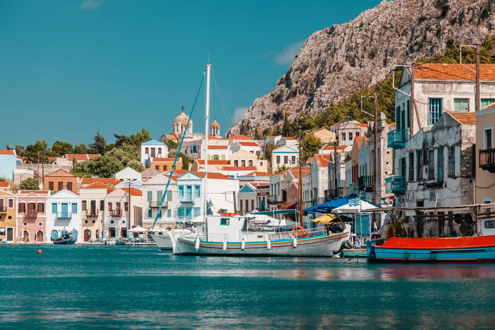 Meis Adası'nın berrak masmavi sularında demirlemiş rengarenk tekneleriyle hareketli limanı, sahili süsleyen pastel tonlarda boyalı geleneksel evleri, tepede yükselen kubbeli kilisesi ve arkasındaki heybetli kayalık dağ manzarasıyla pitoresk bir kasaba görünümü sunuyor.
