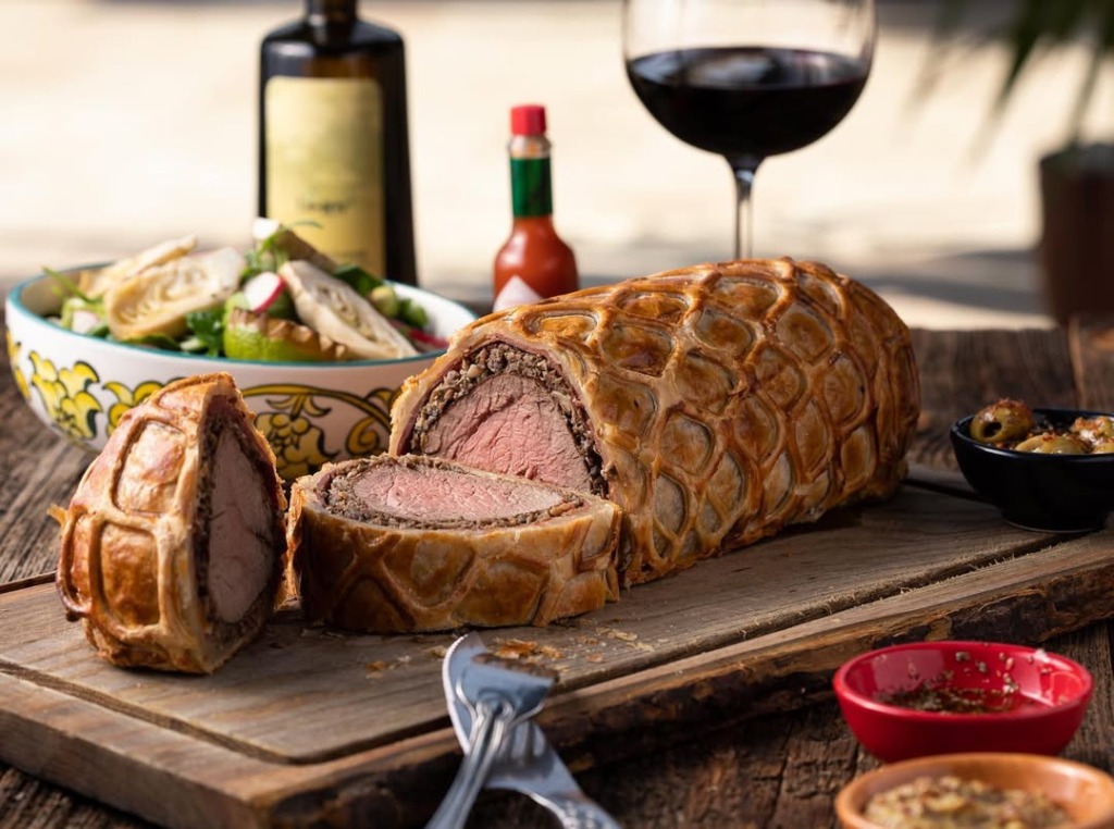 Rustik ahşap bir kesme tahtası üzerinde, altın rengi desenli hamuruyla mükemmel pişmiş pembe et filmini ortaya çıkaran, taze kesilmiş iştah açıcı bir Beef Wellington, yanında renkli salata, bir kadeh kırmızı şarap ve çeşitli soslarla birlikte sunuluyor; bu görüntü Çeşme’nin en iyi happy hour ve gece mekanları'nda yaşanabilecek enfes bir yemek deneyimine işaret ediyor.