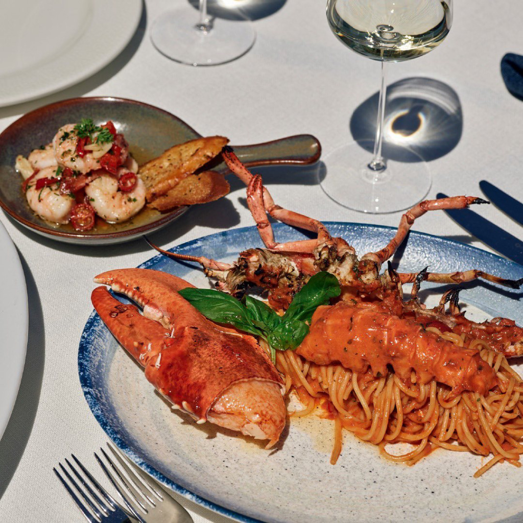 Çeşme ve Alaçatı'nın romantik restoranlarında çiftler için özel olarak hazırlanmış, kremalı domates soslu spagetti üzerinde taze fesleğen yapraklarıyla süslenmiş kocaman bir ıstakoz kıskacı ve gövdesi ile sunulan zarif ana yemek, yanında sarımsaklı karides ve kızarmış ekmeklerin olduğu küçük bir kaseyle, beyaz örtülü bir masada duran kadehlerdeki beyaz şarap eşliğinde iştah açıcı bir şekilde görüntüleniyor.