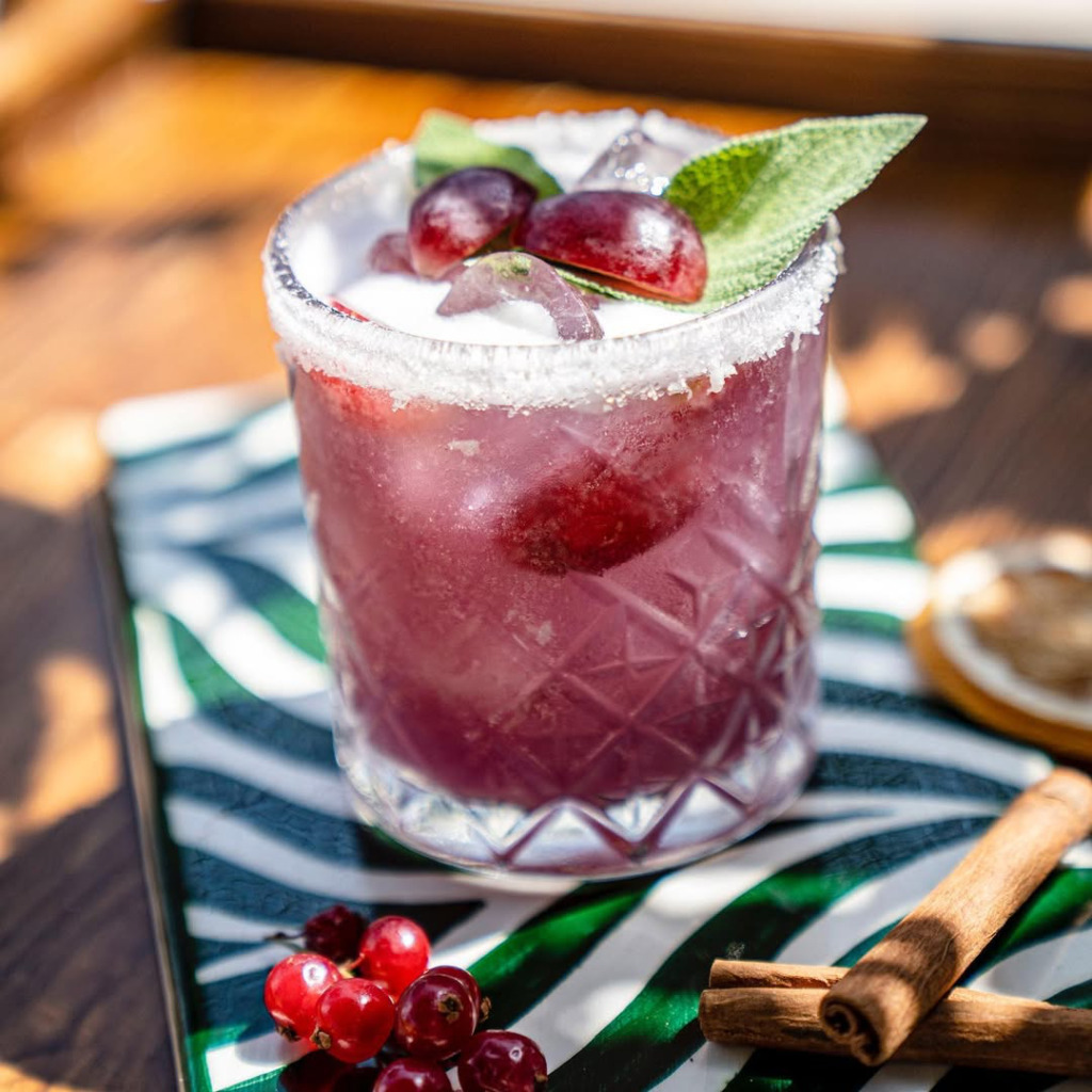 Çeşme'nin en iyi happy hour ve gece mekanlarında sunulan kokteyllerden biri olan, şekerli kenarlı kadehte, mor renkli, ferahlatıcı ve buzlu içecek; üzerinde kırmızı üzümler ve yeşil nane yaprakları ile süslenmiş, yanında taze kızılcıklar ve tarçın çubukları bulunan bu özel sunum, Alaçatı'daki eğlenceli anlara davet ediyor.