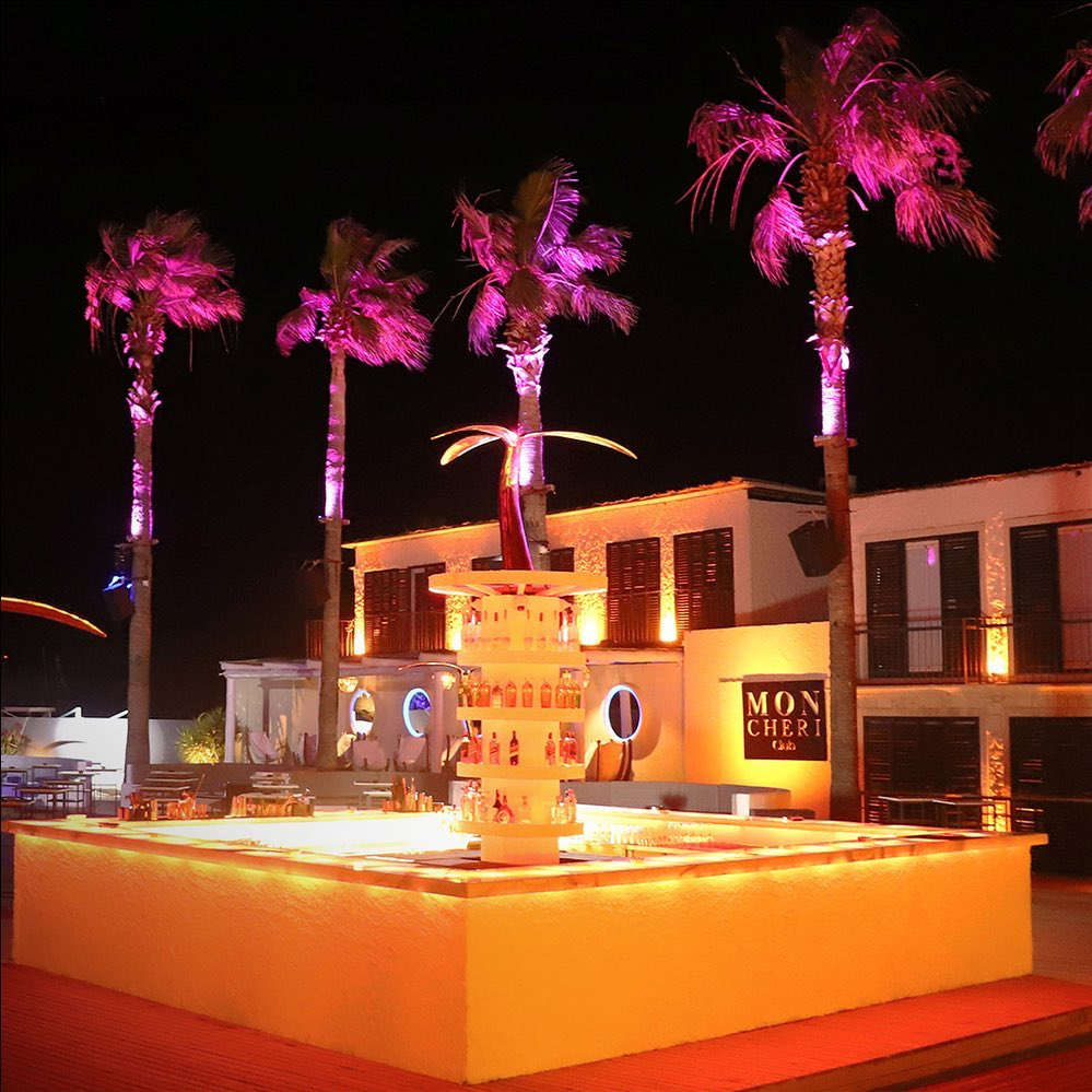 Alaçatı'daki Mon Cheri gece kulübünde, pembe ışıklarla aydınlatılmış uzun palmiyelerin altında, içecek şişeleriyle dolu çok katlı barın ve ışıklı bar tezgahının bulunduğu hareketli bir happy hour ortamı geceye hazırlanıyor.