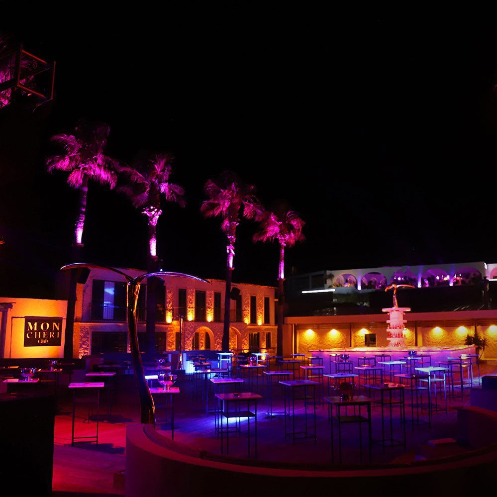 Mon Cheri Beach Club’ın geceye özel mor ve pembe aydınlatmalı palmiyelerle çevrili, mavi ve kırmızı tonlarda ambiyans ışıklarının yansıdığı, çok sayıda masanın hazırlandığı açık alanı, Çeşme’nin hareketli happy hour ve gece mekanları atmosferini yansıtıyor.