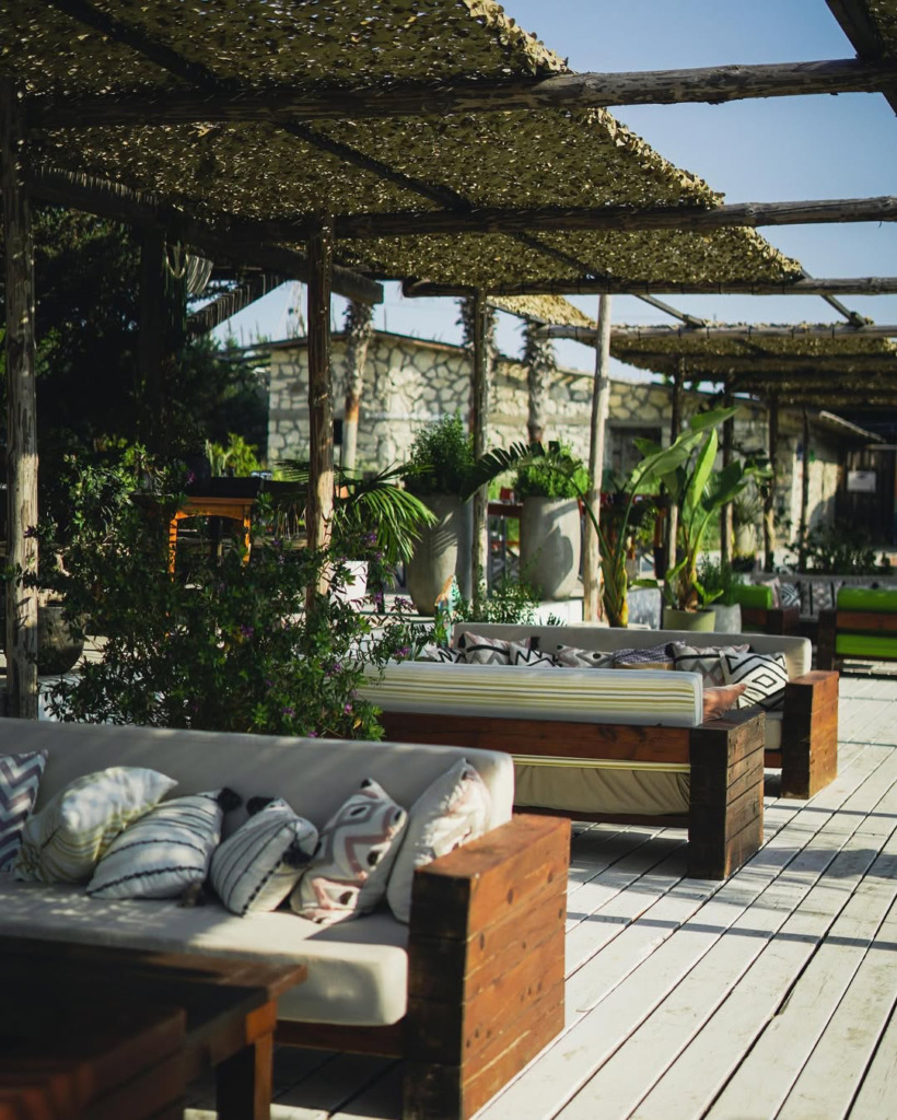 Çeşme’nin hareketli happy hour ve gece mekanları için ideal, boho-şık bir atmosfer sunan açık hava bir alanda, desenli yastıklarla süslenmiş ahşap banklar ve şık kanopi gölgeleri altında dinlenmeye davet eden yemyeşil bitkilerle çevrili taş yapılar görünüyor.