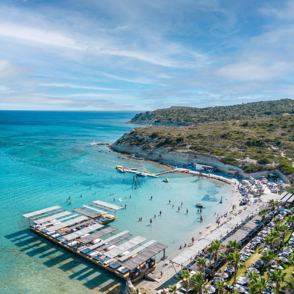 Çeşme'nin en iyi happy hour ve gece mekanlarından biri olan bu canlı plaj kulübünün kuşbakışı görüntüsünde, turkuaz rengi sularında yüzen ve sahildeki şezlonglarda güneşlenen kalabalıklar, gölgelikli yüzen iskeleler, palmiyeler ve çevredeki yemyeşil yamaçlar göz alıyor.