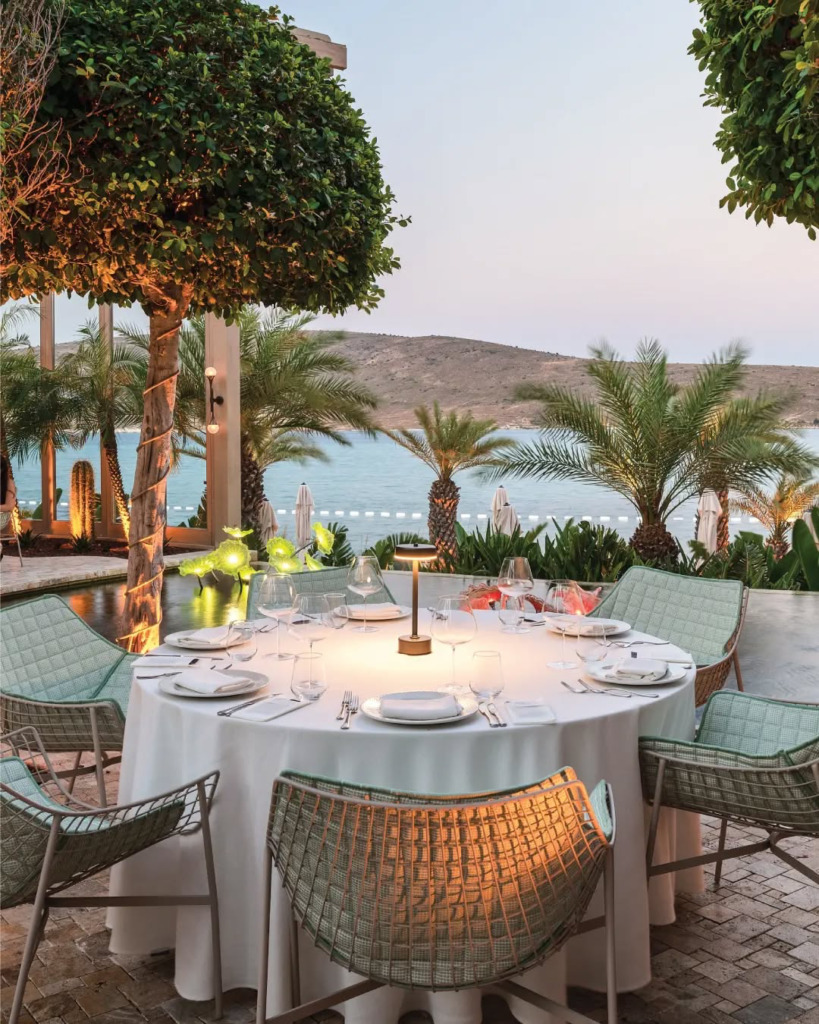 Çeşme/Alaçatı'nın romantik atmosferini yansıtan bu resimde, akşamüzeri deniz manzaralı lüks bir açık hava restoranında beyaz masa örtüsüyle zarifçe hazırlanmış yuvarlak bir yemek masası, etrafında modern tasarımlı sandalyeler ve loş ışıklar dikkat çekerken, fonda palmiye ağaçları ve sakin Ege suları uzanıyor.