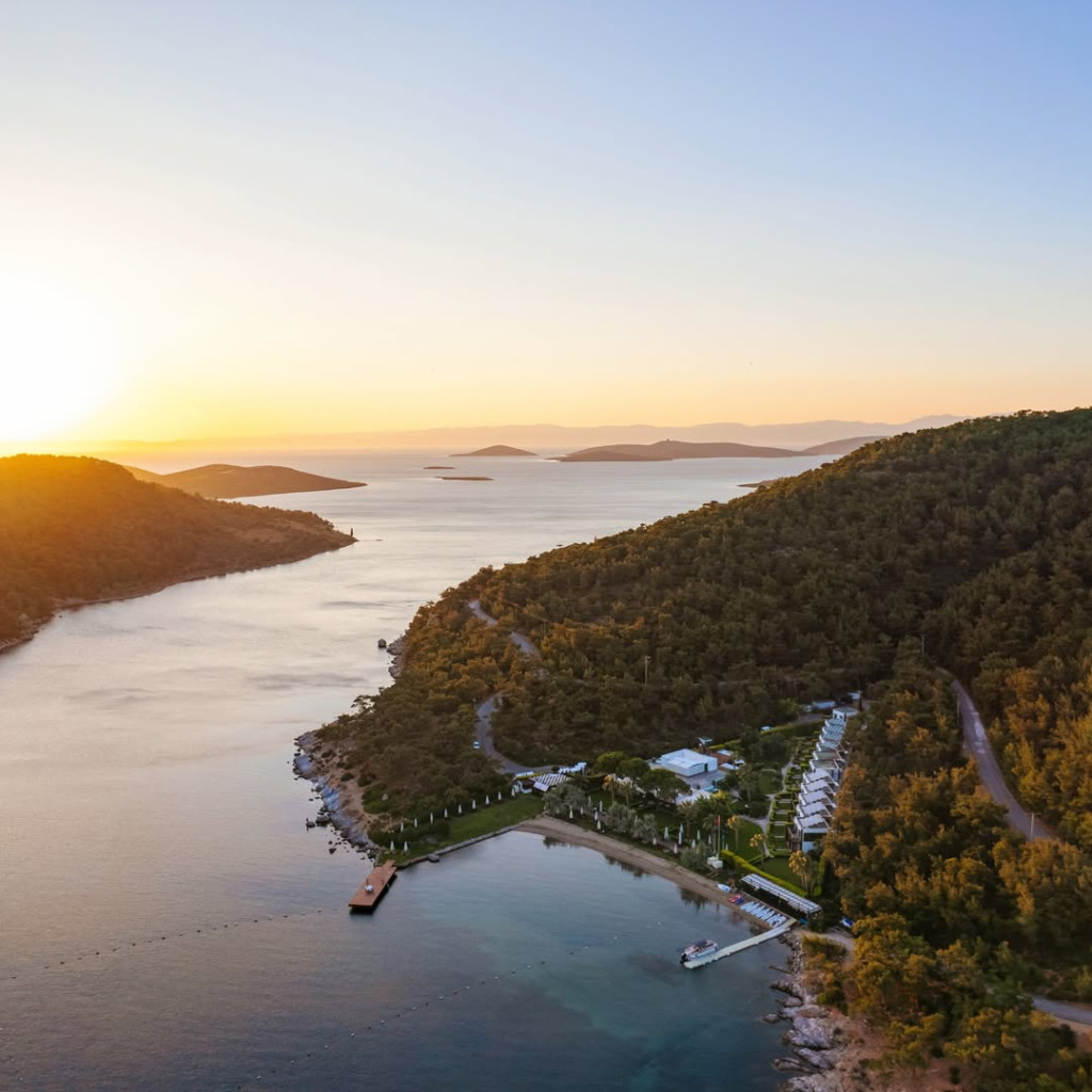 Ayvalık Körfezi'nin yemyeşil tepelerle çevrili bir kıyısında, güneşin batışıyla altın rengine bürünen denize nazır modern yapılar, özel bir plaj ve iskelelerin Cunda Adası seyahat rehberi konseptini yansıtan sakin bir kuşbakışı görüntüsü.