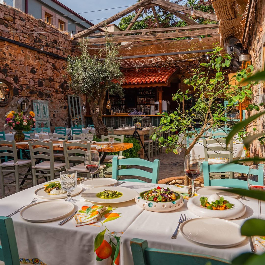 Cunda'nın karakteristiğini yansıtan taş duvarlı bir açık hava restoranında, örtülü masalarda şarap ve Ege mutfağından rafine yemekler servis edilmiş, etraftaki zeytin ağaçları ve açık mavi sandalyelerle birlikte adanın sakin ve zarif ambiyansını taş sokakların ruhuyla birleştiren keyifli bir akşam yemeği manzarası.
