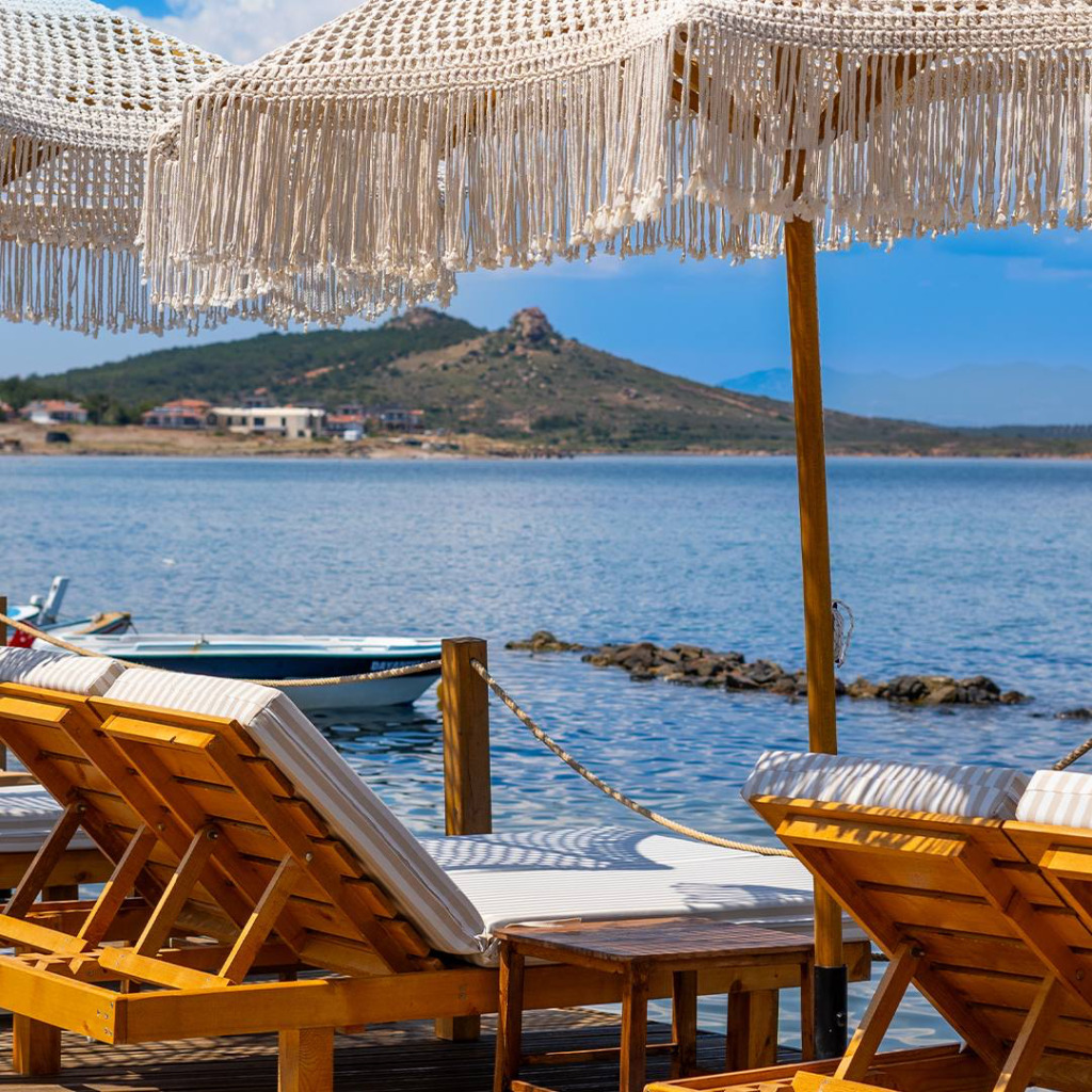Cunda Adası'nda, Ege'nin berrak ve sakin sularına nazır bir ahşap iskelede, püsküllü şemsiyelerle gölgelenmiş rahat şezlonglar, adanın huzurlu atmosferinde keyifli bir mola ve unutulmaz bir seyahat deneyimi sunuyor.