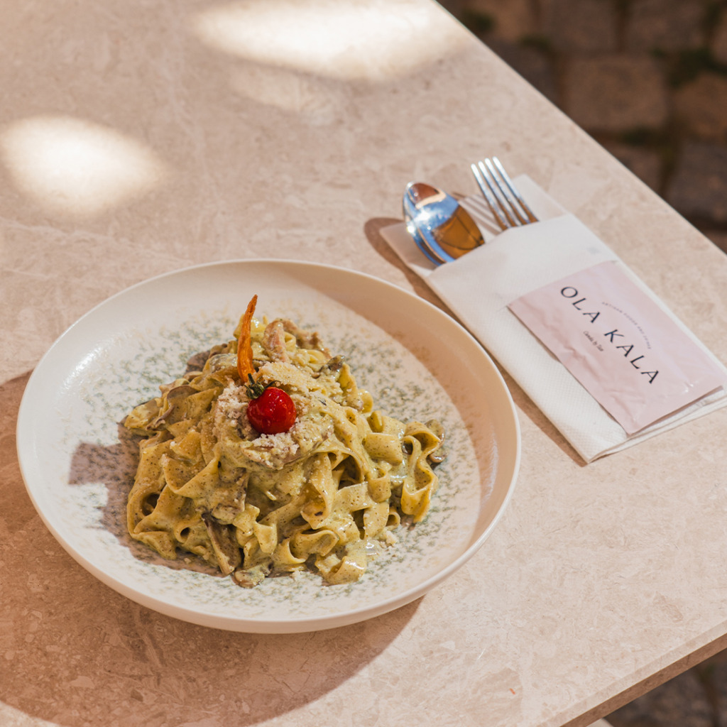 Cunda'nın tarihi dokusunu hissettiren taş sokaklarında, Ola Kala restoranında sunulan, bol kremalı mantar ve çeşitli otlarla zenginleştirilmiş taze fettuccine, üzerine rendelenmiş peynir ve bir kiraz domatesle süslenmiş halde mermer bir masada, markalı peçetesi ve parlayan çatal bıçağıyla güneşli bir öğle yemeği deneyimini gözler önüne seriyor.