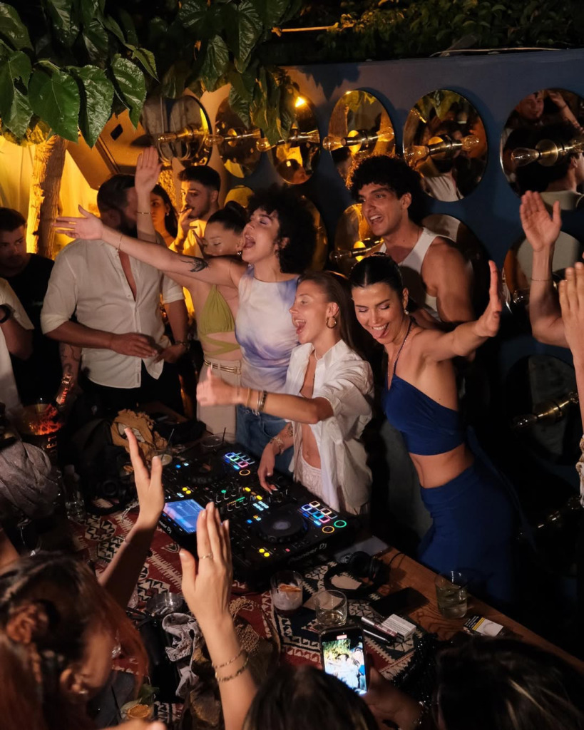 Bir DJ'in başında olduğu, ışıklı konsolun önünde enerjiyle ellerini havaya kaldırıp dans eden kalabalık, Çeşme'nin hareketli gece mekanlarından birinde happy hour sonrası coşkulu bir partinin tadını çıkarıyor.
