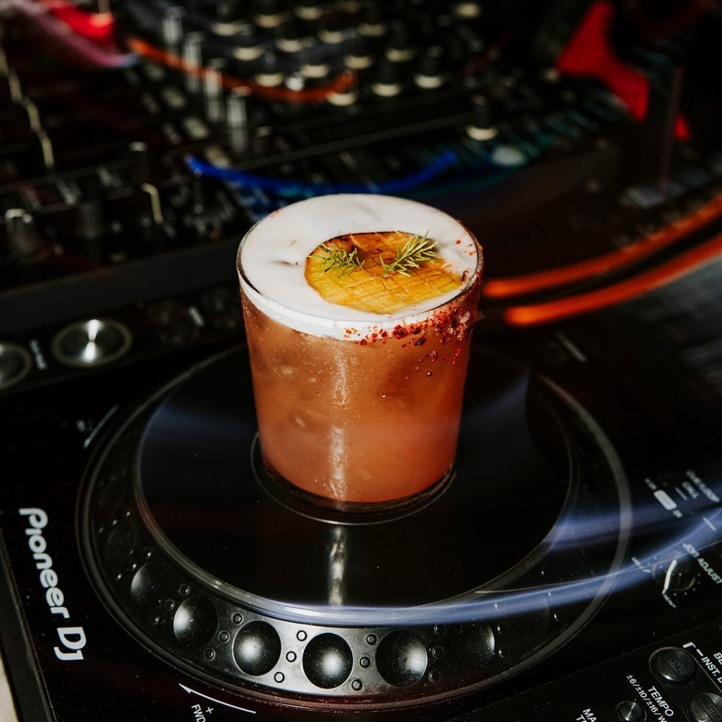 Bir Pioneer DJ pikapının döner tablası üzerinde duran, kenarı kırmızı baharatlarla süslü, üzerinde zengin beyaz köpük ve ızgara ananas dilimiyle taze otlar olan amber renkli bir kokteyl, Çeşme'nin canlı happy hour etkinlikleri ve hareketli gece mekanlarının enerji dolu atmosferini vurguluyor.