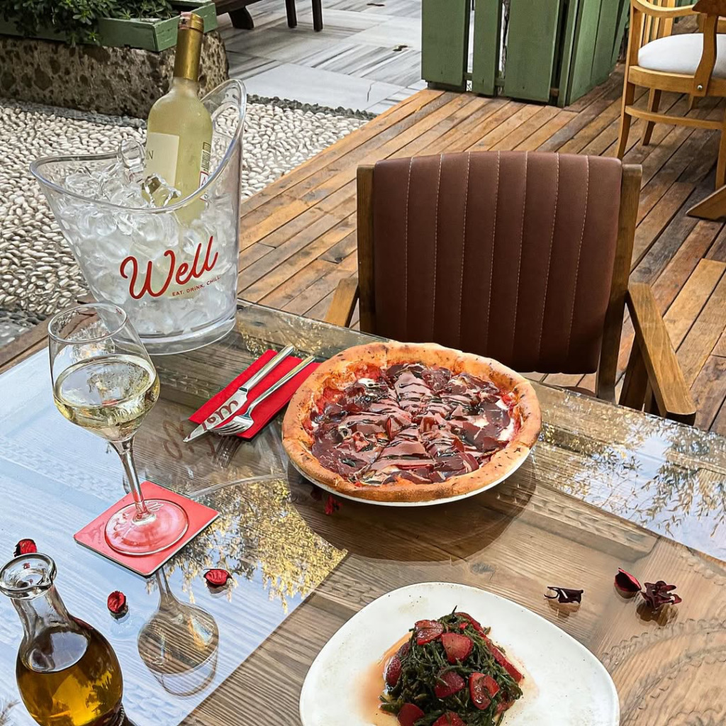 Çeşme'nin hareketli happy hour ve gece mekanlarından biri olan Well'de, markanın logosunu taşıyan buz kovasında soğuk beyaz şarap, taze malzemelerle hazırlanmış pizza ve çilekli yeşillikler salatasının cam masada keyifle sunulduğu şık bir dış mekan düzenlemesi gösteriliyor.