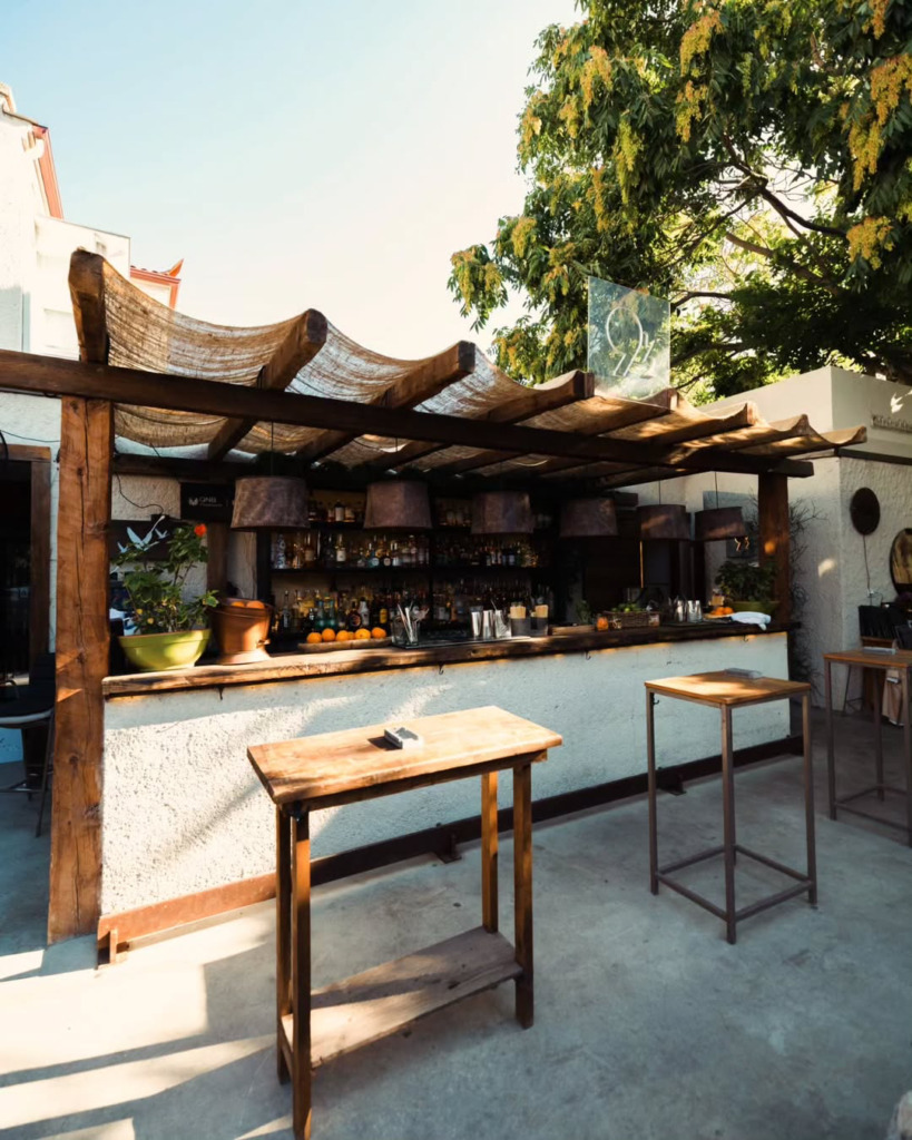 Çeşme’nin en iyi happy hour ve gece mekanlarından biri olan Isla Alaçatı’da, ahşap pergolalı açık bir barda şişeler, taze meyveler ve kokteyl malzemeleri misafirler için hazır beklerken, güneşin vurduğu ahşap masalar ve genel düzenleme, mekanın davetkar ortamını sergiler.