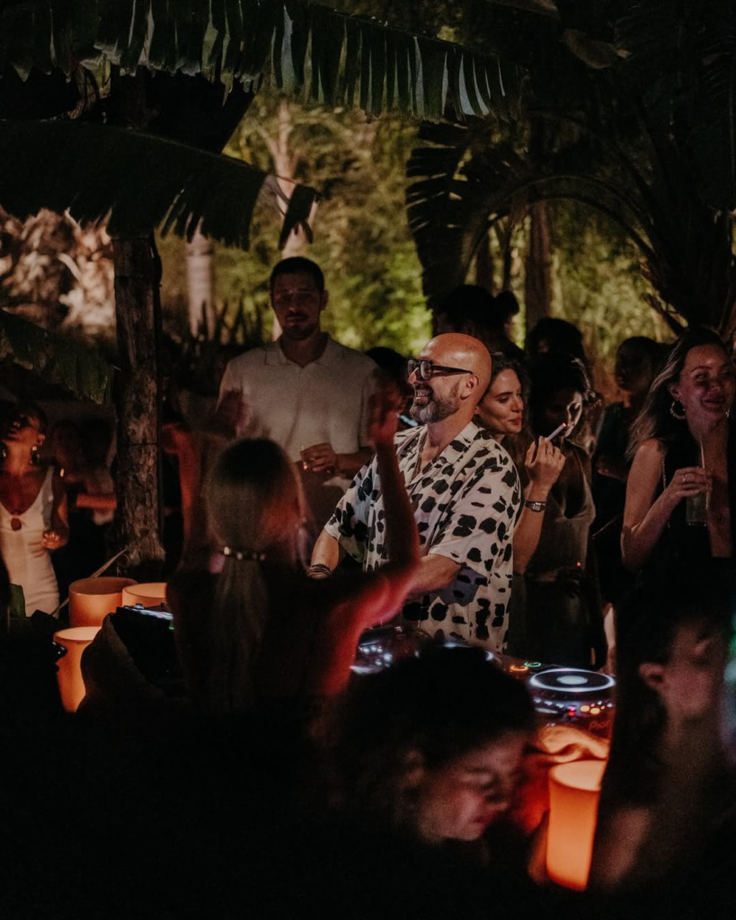 Bir happy hour veya gece mekanında, tropikal bitkilerin arasında loş ışıklar altında, kel ve gözlüklü bir DJ'in coşkulu kalabalığa enerji veren müzikler çalarken etrafındaki misafirlerin dans edip içkileriyle eğlendiği hareketli bir an.