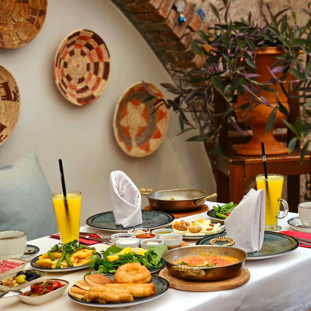 Cunda seyahat rehberi içerisindeki butik otellerden birinde, adanın ruhunu yansıtan taş duvarlı ve geleneksel sepetlerle dekore edilmiş bir mekanda, portakal suyu, menemen, pişi, çeşitli peynir ve zeytin gibi Ege'ye özgü zengin lezzetlerle dolu, güne başlangıç için davetkar bir kahvaltı sofrası.