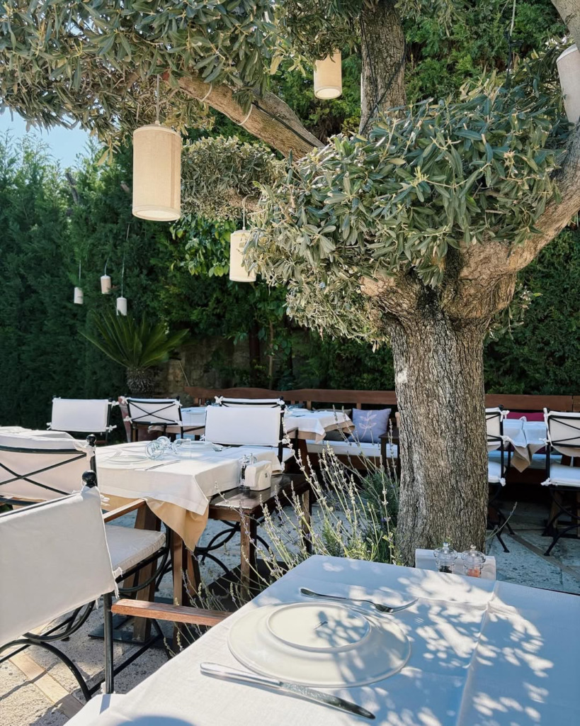 Çeşme/Alaçatı'nın romantik atmosferini yansıtan bu açık hava restoranında, beyaz örtülü zarif masalar büyük bir ağacın gölgesine serilmiş, dallarından sarkan fenerler ve çevresindeki yeşilliklerle huzurlu bir yaz akşamı yemeği için ideal bir ortam sunuluyor.