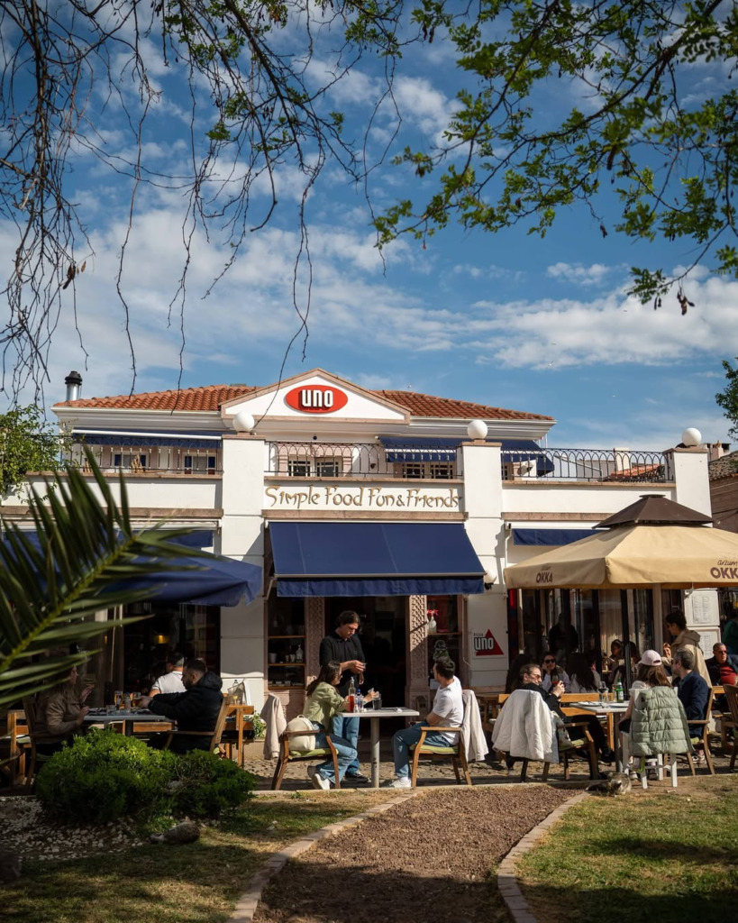 Cunda Adası'nın merkezinde yer alan, UNO tabelalı beyaz renkli binası ve mavi tente altındaki kalabalık dış oturma alanıyla Uno Pizza, taş sokakların enerjisini yansıtan keyifli bir Ege akşamı sunuyor.