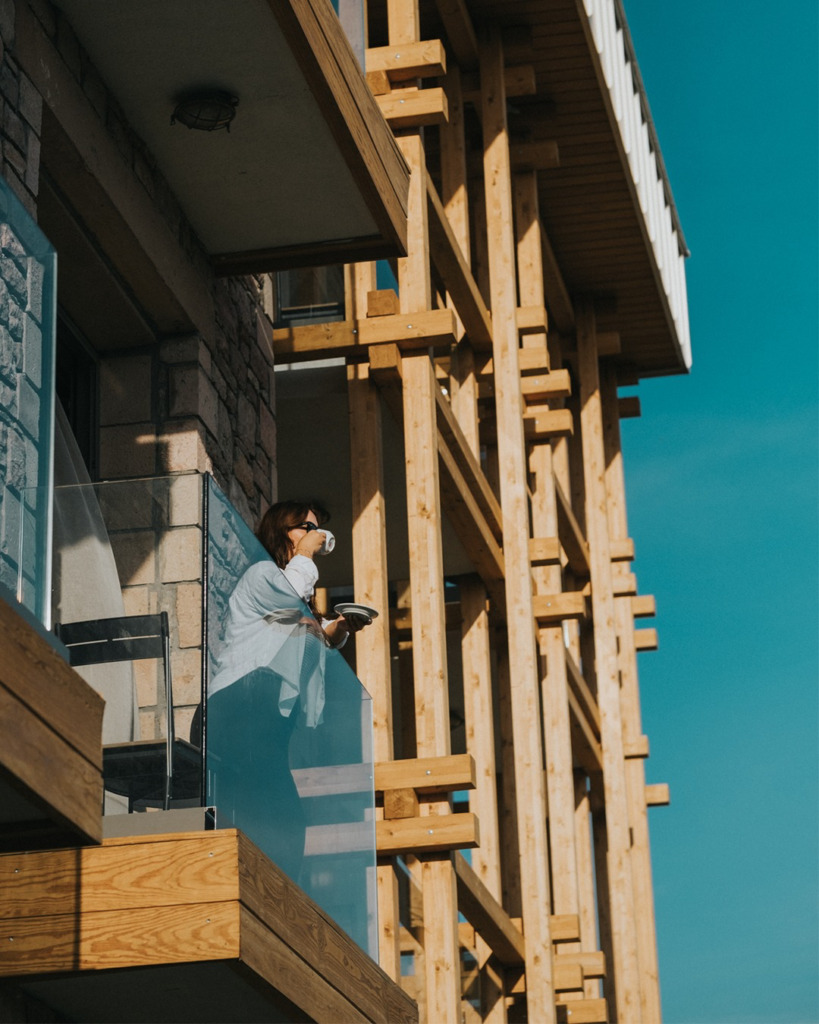 Güneşli bir havada, Cunda'nın modern bir butik otelinin taş duvarlı ve ahşap detaylı balkonunda elinde fincanıyla dışarıyı seyreden gözlüklü bir kadın, adanın huzurlu ambiyansının ve özgün mimarisinin keyfini çıkarıyor.