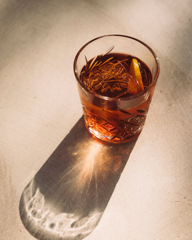 Bir happy hour veya gece mekanında, yüzeyde uzun gölgesi ve ışık yansımalarıyla dikkat çeken, içinde büyük desenli buz ve limon dilimi bulunan, karmaşık kesme cam işlemeli şık bir kokteyl, Çeşme'nin hareketli atmosferini yansıtıyor.