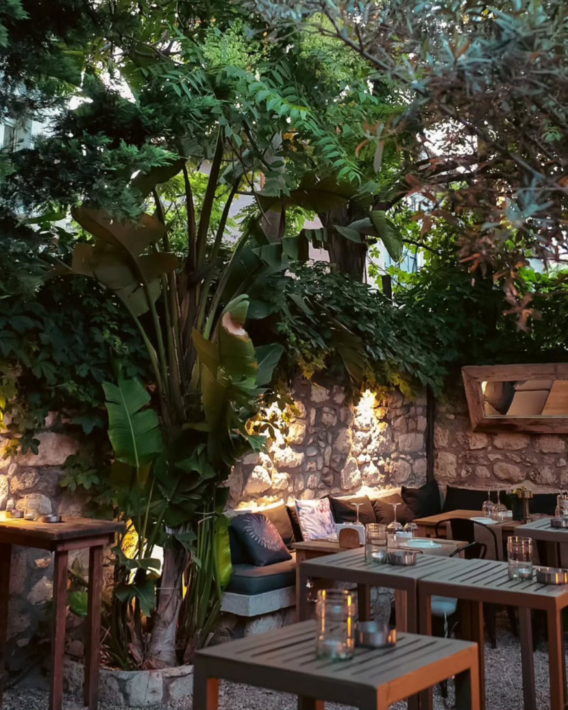 Çeşme'nin hareketli gece mekanlarından Isla Alaçatı'nın yemyeşil tropikal bitkilerle bezenmiş, taş duvarlı dış oturma alanı, loş ışıklı ve happy hour için özenle dizilmiş ahşap masalarıyla davetkar bir atmosfer sunuyor.