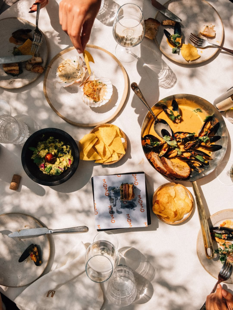 Bir kişinin deniz tarağı tabağından bir cips aldığı, midye, guacamole ve diğer lezzetlerin sunulduğu, beyaz örtülü bir masada güneş ışıkları altında keyifli bir happy hour yemeği, Çeşme'nin en iyi mekanlarından The Beach of Momo'nun atmosferini yansıtıyor.