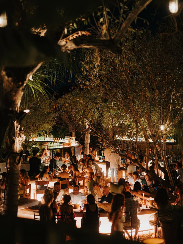 Çeşme'nin hareketli gece hayatından bir kesit sunan bu fotoğrafta, tropikal ağaçlar ve loş ışıklandırmalar altında, merkezi bir bar etrafında toplanmış kalabalık bir grup insan, kokteyllerini yudumlayarak sosyalleşiyor ve keyifli bir happy hour deneyimi yaşıyor.