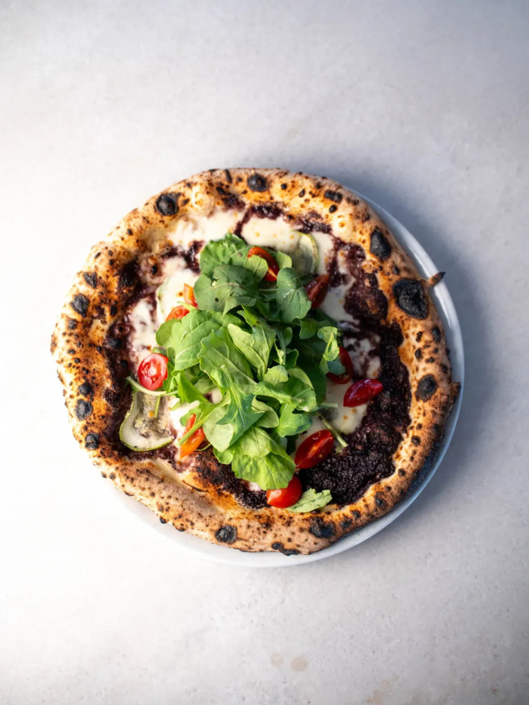 Cunda Seyahat Rehberi kapsamında adanın gastronomik zenginliğini yansıtan, kenarları nar gibi kızarmış, közlenmiş Napoli usulü pizzanın üzerinde bolca taze roka, parlak kırmızı kiraz domatesler ve erimiş peynirle harmanlanmış koyu renkli sos, Cunda'nın taş sokaklarında sunulan enfes lezzetlerini gözler önüne seriyor.