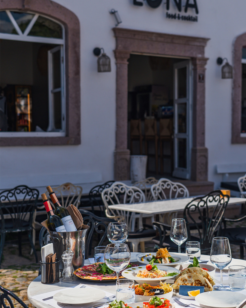 Cunda'nın taş sokaklarından birinde yer alan Luna Cunda restoranının dış mekanında, üzerinde ince hamur pizza, karidesli makarna, taco ve bir şarap kovasıyla çeşitli Ege lezzetleriyle dolu şık bir masa, akşam yemeği için hazır bekliyor.